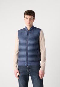 Tumši zils pārklātais pūkains veste ar rāvējslēdzēju priekšpusē, ribotām apkaklēm un jostu; uzvilkta virs krēmkrāsas garām piedurknēm krekla.