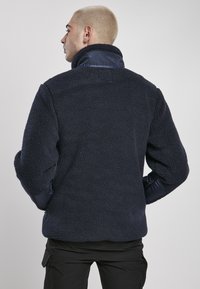 Tumesinine fliisk jacket pehme ja tekstureeritud viimistlusega; kõrgema kaelusega ja kontrastsete siledate kanga aksentidega varrukatel.