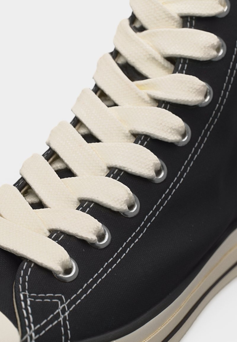 Primo piano di larghe stringhe color crema infilate attraverso occhielli metallici su una sneaker in tela nera con cuciture bianche.