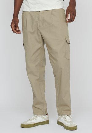Beige cargo nadrág oldalzipzáros zsebekkel, elasztikus mandzsettákkal és laza szabással, fehér és olíva zöld sportcipőkkel párosítva.
