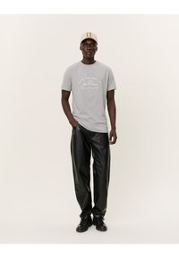 Camiseta de algodón gris con texto gráfico en blanco, combinada con pantalones de cuero negros y zapatillas negras. El modelo lleva una gorra beige con logo.