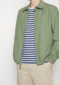 Veste légère vert olive avec un col, fermeture éclair et poignets élastiques sur une chemise rayée bleue et blanche.