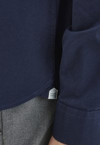 Navy kék pamut hosszú ujjú ing lekerekített aljjal és kontrasztos varrással, amelyen a "Casual Friday" feliratú címke található. Texturált anyag.