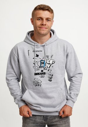 Ung mand smiler, iført en lysegrå hættetrøje med sorte og blå grafiske designs og tekst, stående foran en ensfarvet hvid baggrund.