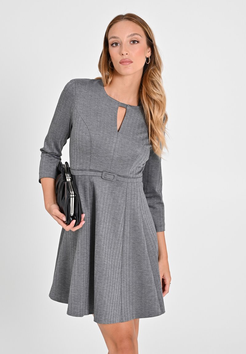 Graues Kleid mit Dreiviertelärmeln, Schlüsselloch-Ausschnitt, taillierter Taille, weit schwingendem Rock und vertikaler Rippstruktur. Hält eine schwarze Clutch.