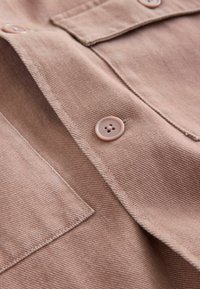 Gros plan sur une chemise en tissu beige avec des coutures visibles, un bouton et une poche poitrine, montrant une texture et des détails fins.