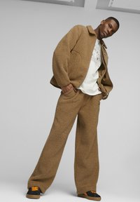 Completo felpa marrone e morbida con giacca e pantaloni a vestibilità rilassata. L'outfit include una maglietta bianca con grafica e sneakers nere con dettagli arancioni.