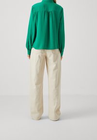 Blusa verde con maniche lunghe e schiena arricciata, abbinata a pantaloni beige a gamba larga. Il tessuto sembra liscio e leggero.