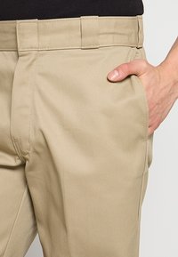 Main dans la poche droite d'un pantalon beige avec passants de ceinture sur une personne portant une chemise noire.