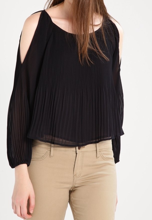 Femme portant un chemisier noir plissé à épaules dénudées avec manches longues et un pantalon beige ajusté, sur un fond uni.