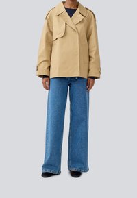 Beige dobbeltradet jakke med skulderlameller, parret med brede blå denimjeans. Stoffet er glat med en struktureret pasform.