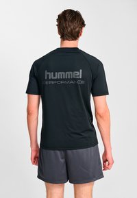 Svart atletisk t-shirt med en slät yta, med "hummel PERFORMANCE" tryckt i fet grå stil på baksidan, kombinerad med grå shorts.