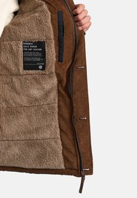 Ruskea corduroy-takki, jossa on pehmeä beige fleece-vuori. Varustettu vetoketjuilla ja hoitolapulla sisäpuolella. Kestävä suunnittelu kaikkiin vuodenaikoihin.