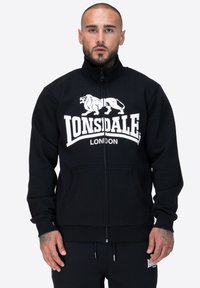 Czarna bluza z zamkiem wykonaną z materiału z prążkowanymi mankietami i pasem. Na przodzie znajduje się biały logo lwa oraz tekst "Lonsdale London".