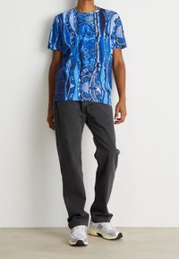 Person som bär en blåmönstrad kortärmad skjorta, mörkgrå raka jeans och vita New Balance-sneakers som står mot en enfärgad vägg.