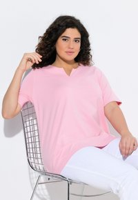 Tunic rosa chiaro a maniche corte con scollatura a V e una vestibilità comoda, abbinata a pantaloni bianchi. Il tessuto appare liscio e morbido al tatto.