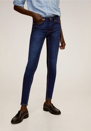 Persoon draagt donkerblauwe skinny jeans, lichtblauw opgerold overhemd en zwarte loafers, staand met één hand in de broekzak.