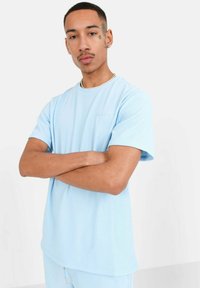 Noor mees, kelle käed on risti ja seljas on helesinine ribakujulise mustriga T-särk ja vastavad püksid, seistes ühtlase heleda tausta ees.