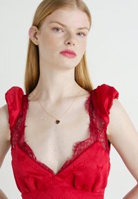 Rode jurk met kantaccenten op de schouders en de halslijn, textuurstof met bloemmotieven en een aansluitende silhouet. Gouden kettingaccent.