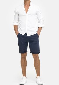 Chemise boutonnée en lin blanc, shorts bleu marine retroussés, baskets blanches. La chemise a un col classique, et les shorts possèdent des poches latérales.