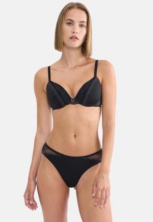 Donna che indossa un completo di lingerie in pizzo nero con spalline regolabili, in piedi davanti a uno sfondo bianco semplice con un'espressione neutra.