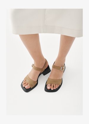 Nohy v béžových semišových sandálech s otevřenou špičkou, kotníkovými pásky a nízkými hranatými podpatky, doplněné světle zbarvenými zkrácenými kalhotami.