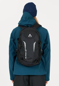 Svart ryggsäck med justerbara remmar, featuring en strukturerad yta, grå detaljer och en logotyp. Bärs över en blå jacka mot en vit bakgrund.