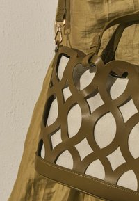 Sac à main beige et olive avec découpes géométriques en cuir tressé, suspendu à l'épaule sur un fond en tissu olive assorti.