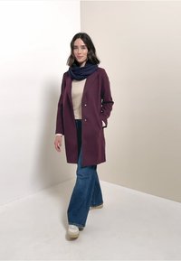 Bordeaux jas met een kraag en twee knopen, gecombineerd met een crèmekleurige trui en blauwe broek met een flared model. Een marineblauwe sjaal zorgt voor kleurcontrast.