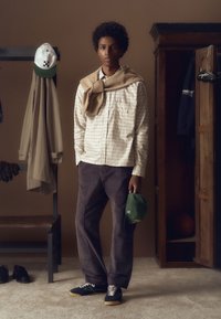 Jeune homme portant une chemise à carreaux et un pantalon foncé tient une casquette verte, debout dans une pièce avec un banc en bois, un porte-manteau et une armoire ouverte.