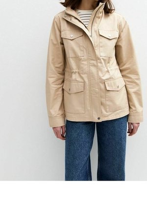 Chaqueta beige con cremallera frontal y bolsillos con botones automáticos, llevada sobre una camisa a rayas, combinada con vaqueros azules en una persona de pie frente a un fondo liso.