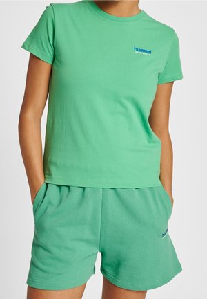 Persona con camiseta deportiva hummel verde y pantalones cortos a juego con las manos en los bolsillos sobre un fondo liso.