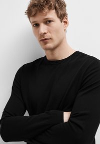 Camisa preta de manga longa feita de tecido suave, com decote redondo e punhos canelados, usada de forma casual com os braços cruzados contra um fundo simples.