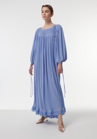 Vestido maxi plisado de color azul claro con un corpiño de fruncido, detalles de cordón y dobladillo de encaje. Combinado con zapatos de tacón transparente.