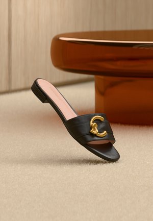 Czarna skórzana sandałka na płaskiej podeszwie z odkrytymi palcami i złotym podwójnym logo C, ustawiona na beżowym dywanie obok zaokrąglonego brązowego drewnianego stolika.