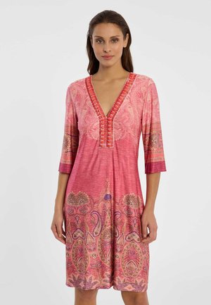 Frau steht und trägt ein knielanges Kleid mit rosa Paisley-Muster, dreiviertellangen Ärmeln und V-Ausschnitt mit Knopfdetails.