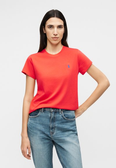 Γυναίκα με μακριά σκούρα μαλλιά, φορώντας φωτεινό κόκκινο κοντομάνικο μπλουζάκι και μπλε τζιν, στέκεται με το ένα χέρι στη μέση.