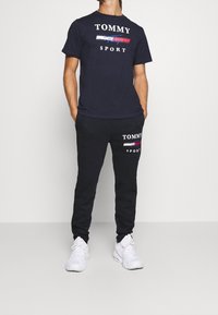 Navy Baumwoll-T-Shirt mit "TOMMY SPORT"-Logo in Weiß und Rot. Schwarze Jogginghosen mit Logo-Druck. Getragen mit weißen Sportschuhen.