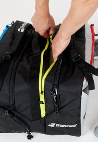 Mani che aprono una borsa sportiva nera Babolat con una cerniera gialla brillante su uno sfondo bianco.