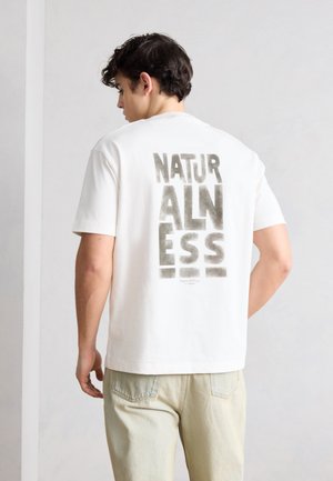 Tânăr purtând un tricou alb cu text mare în blocuri „NATURALNESS” imprimat vertical pe spate.