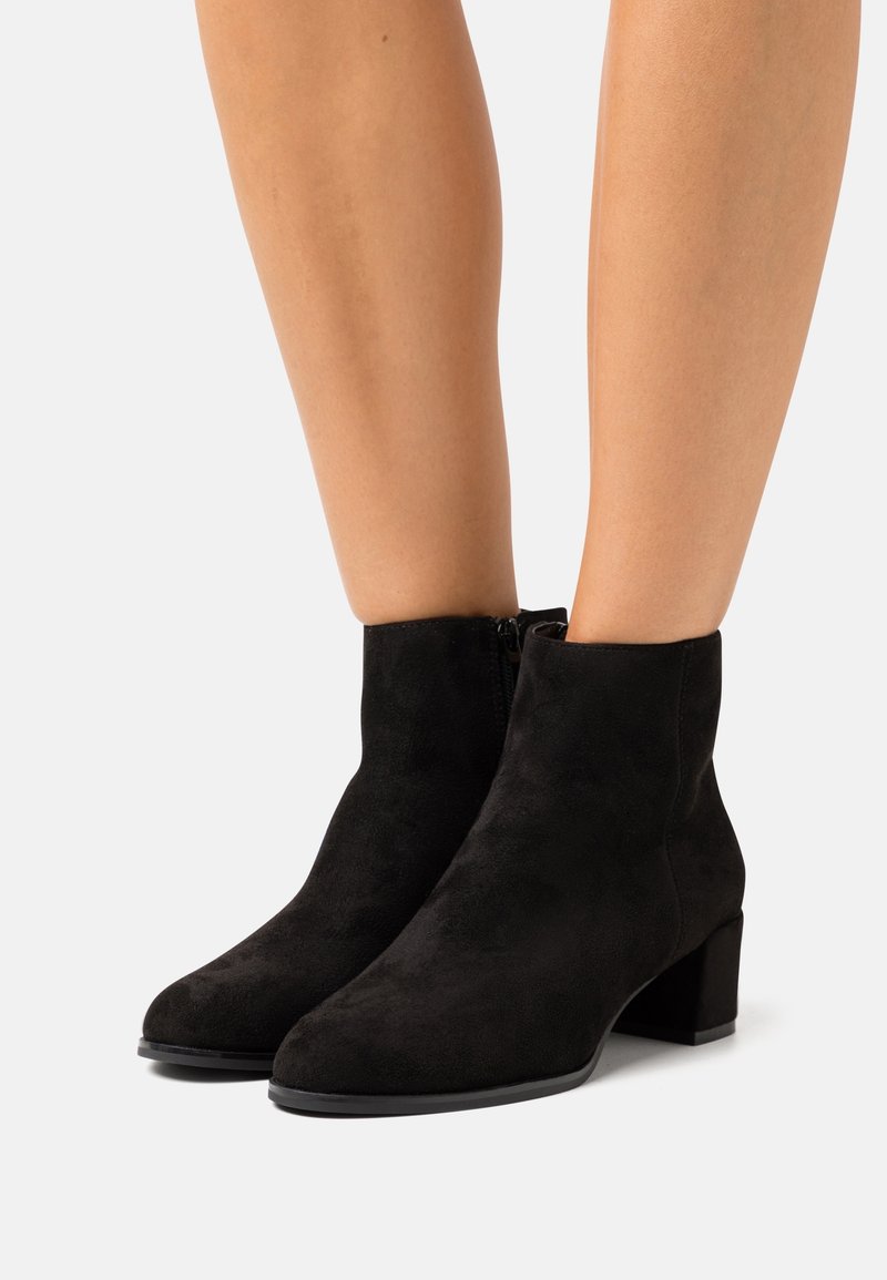 Jambes portant des bottines en daim noir à talons carrés et fermetures éclair latérales sur fond blanc uni.