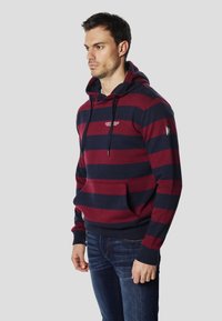 Červená a tmavomodrá pásikavá mikina s kapucňou, predným vreckom, šnúrkou na kapucni a detailom loga na hrudi. Mäkká látka s voľným strihom.