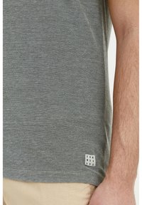 Tricou gri cu maneci scurte dintr-un material texturat, având o etichetă mică cu "DNM WORK KRS" la margine, asortat cu pantaloni bej.