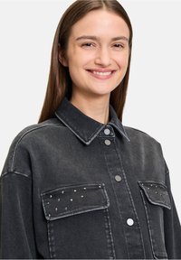 Czarna dżinsowa kurtka z zapięciem na guziki z przodu, dwiema kieszeniami na piersi oraz srebrnymi ćwiekami na klapach kieszeni. Posiada klasyczny kołnierz i luźny krój.