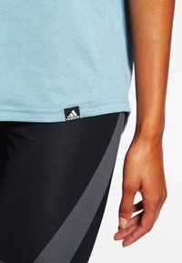 Lichtblauw katoenen T-shirt met een zwart Adidas-logo, gecombineerd met zwarte leggings met grijze diagonale accenten. Soepele textuur.