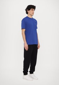 Μπλε βαμβακερό T-shirt με κοντά μανίκια, συνδυασμένο με μαύρες φόρμες και λευκά αθλητικά παπούτσια. Απλός σχεδιασμός χωρίς έντονα μοτίβα ή αξεσουάρ.