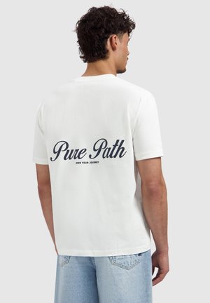 Junger Mann mit lockigem Haar, der ein weißes T-Shirt mit dem schwarzen Schriftzug „Pure Path Own Your Journey“ auf dem Rücken trägt, kombiniert mit hellblauen Jeans.