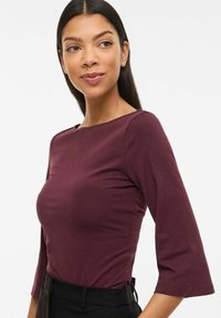 Sieviete ar gariem tumšiem matiem, tērpusies pieguļošā bordo krāsas 3/4 piedurkņu topā un melnās biksēs, skatās nedaudz pa labi ar vieglu smaidu.