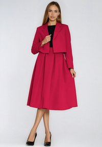 Zestaw z fuksjową marynarką i sukienką z dopasowanym gorsetem, plisowaną spódnicą i krótką marynarką. Czarne grochy zdobią sukienkę.