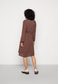 Brun långärmad klänning med åtstramad midja, knälång nederkant och ett luftigt material. Till detta vit sandaler på en grå bakgrund.